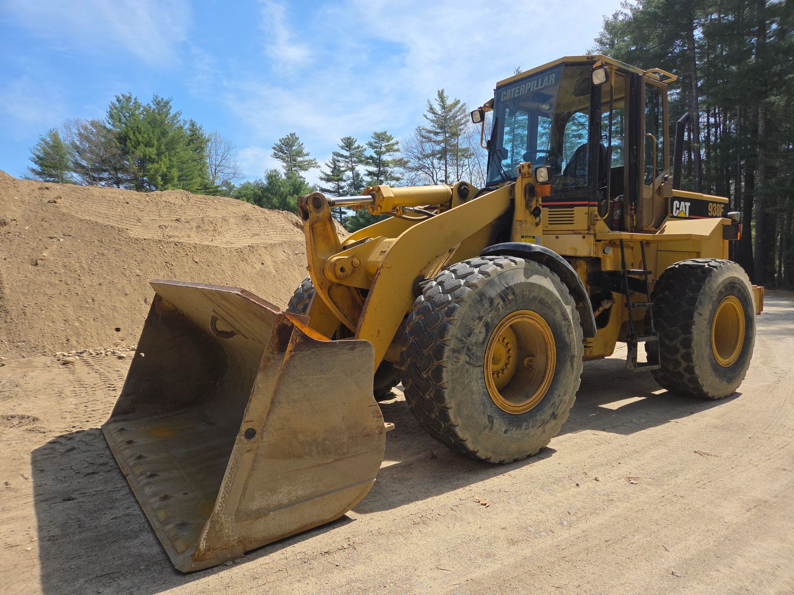 Yellow Caterpillar 938F front loader with a dirt pile and trees in the background.