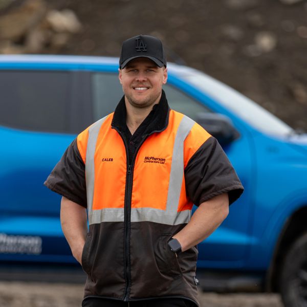 Man in orange and black safety jacket stands in front of blue truck.