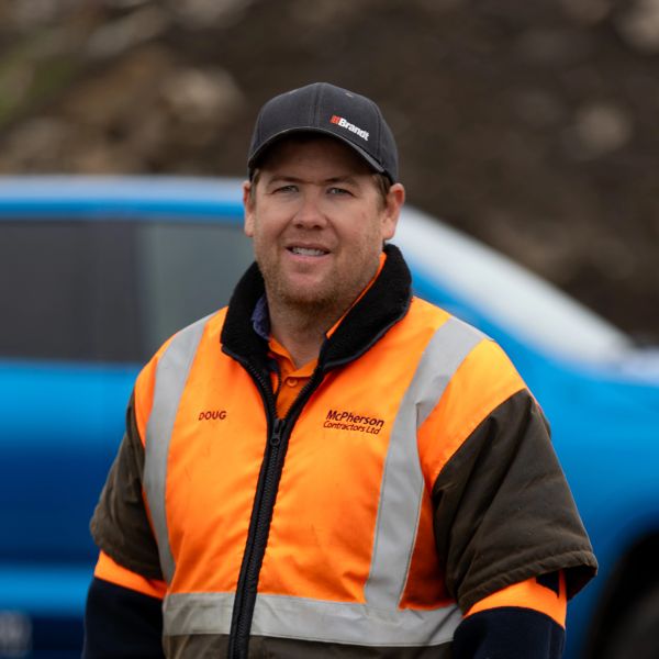 Man in an orange safety jacket and black cap stands outdoors.