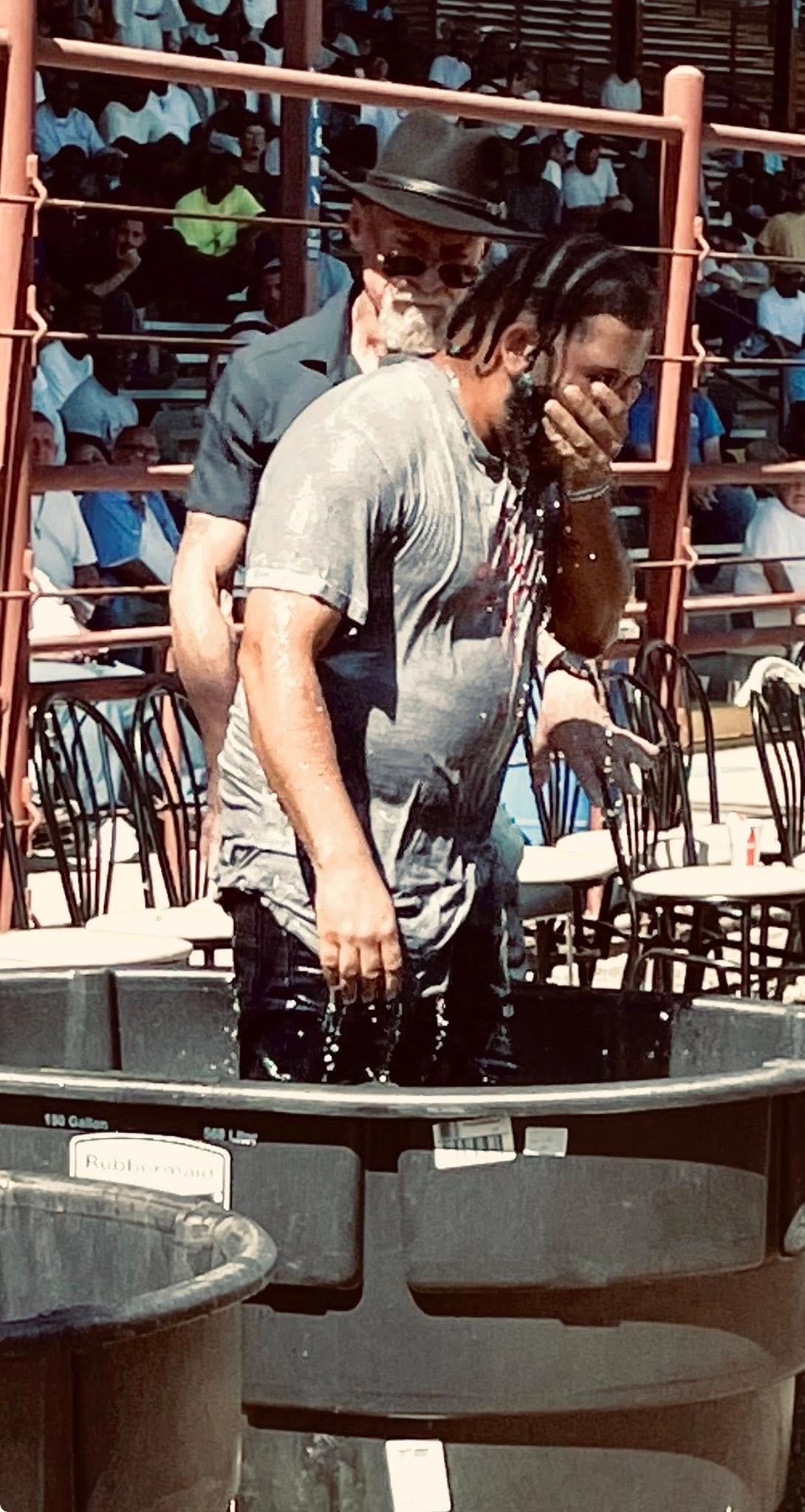 A man emerges from water, covering his face, with a crowd watching behind.