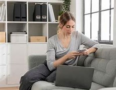 Young woman using smartphone and laptop on sofa in bright room.