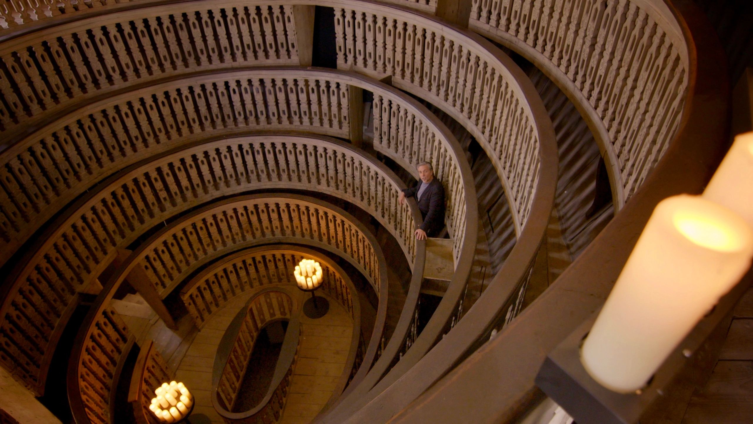 A man stands on a spiraling staircase surrounded by glowing candles.