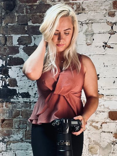Blonde woman with camera stands against a rustic brick wall.