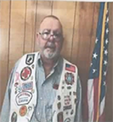 Man with glasses and beard wearing a vest with patches next to an American flag.