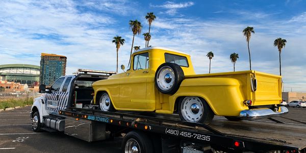 Yellow classic pickup truck on a tow truck under palm trees.