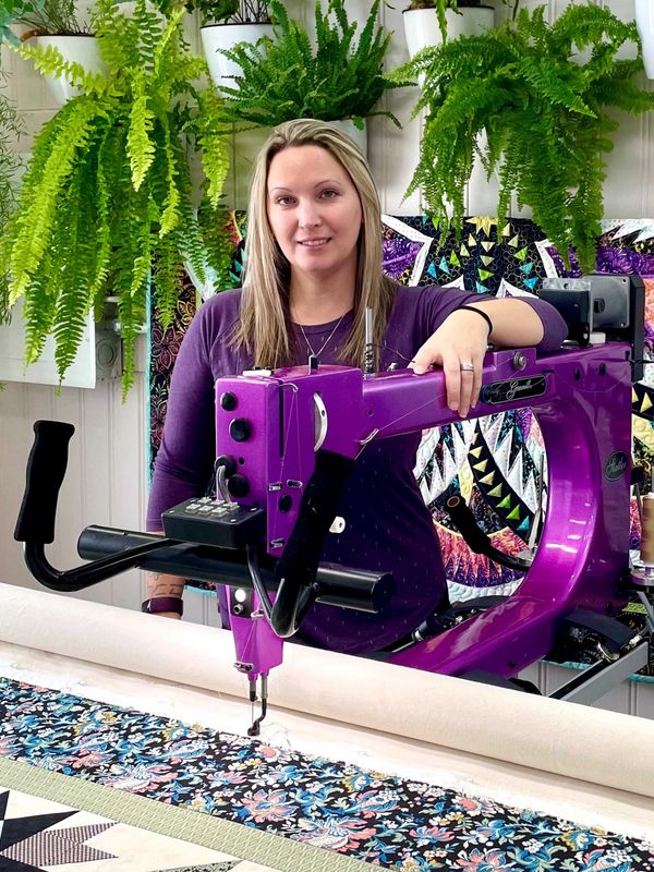 Woman standing behind a purple quilting machine with plants above.