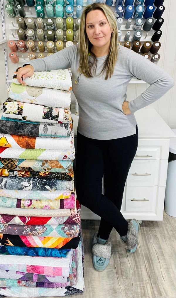 Woman posing with a stack of colorful quilts in a sewing room.