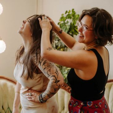 Two smiling women share a joyful moment indoors, one fixing the other's hair.