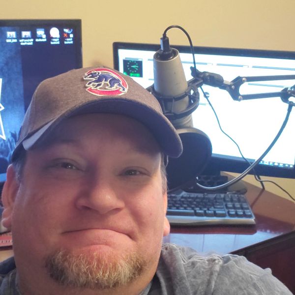 Man wearing a cap with a microphone and computer setup behind him.
