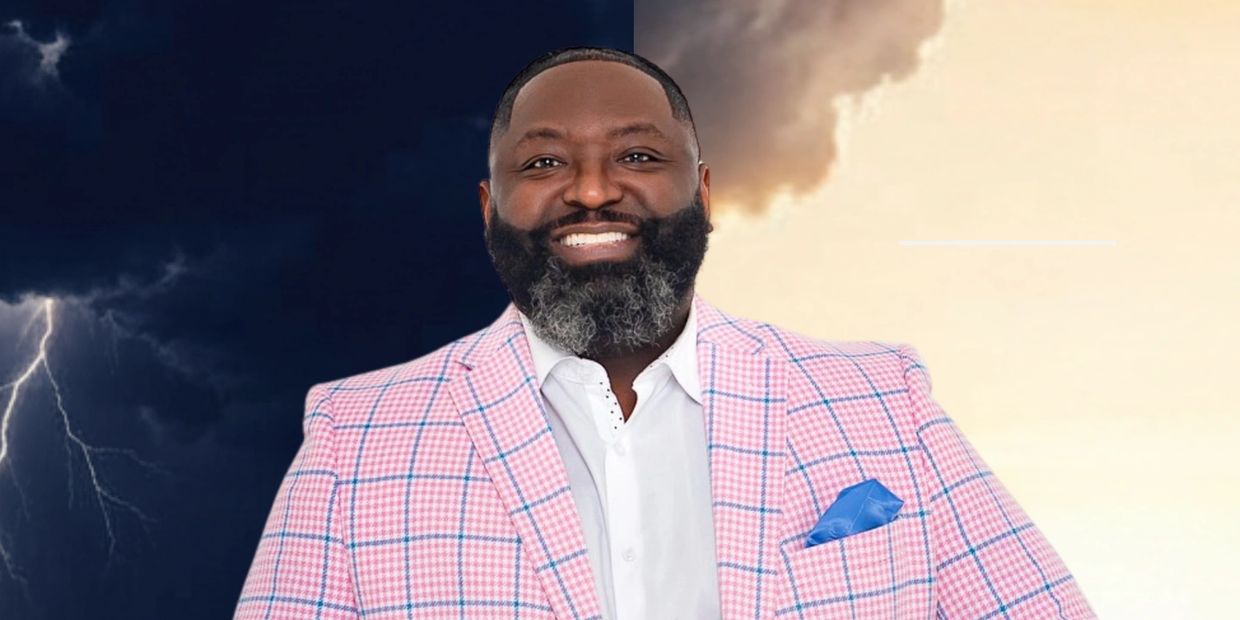 Man smiling confidently in a pink checkered suit against a stormy and sunny background.