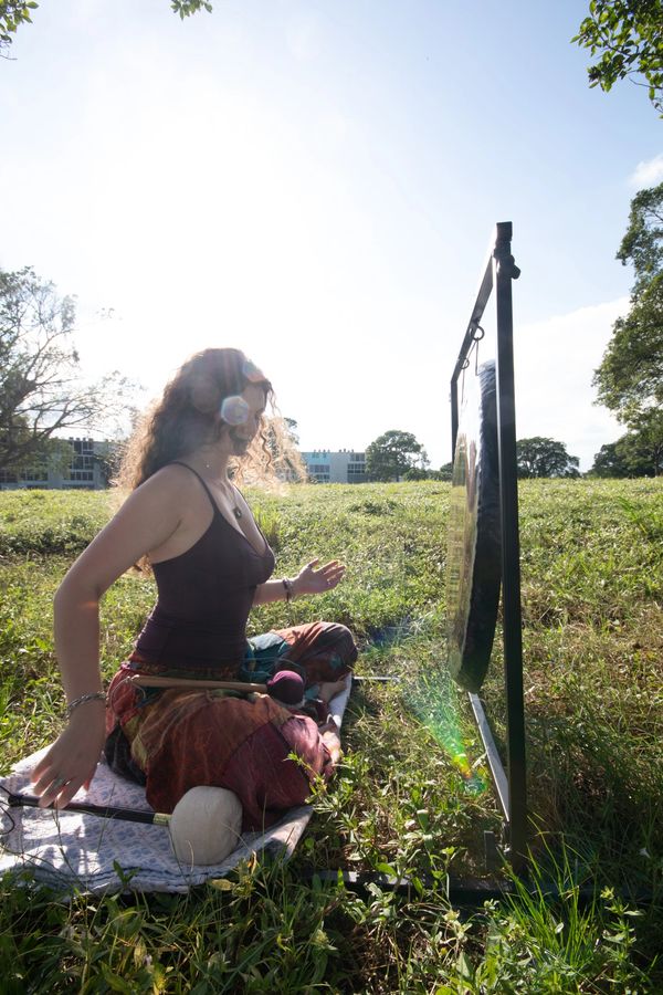 A woman meditatively playing a gong outdoors in sunlight.