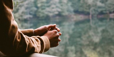Person's hands clasped, resting near a calm lake with blurred trees in the background.