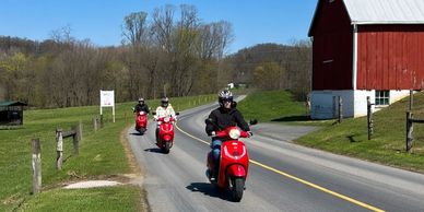 Three people riding red scooters on a rural road near a red barn.