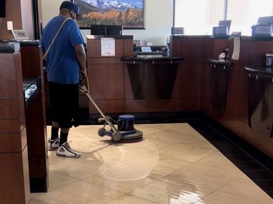 A worker cleaning a tiled floor with a floor buffer in a bank lobby.