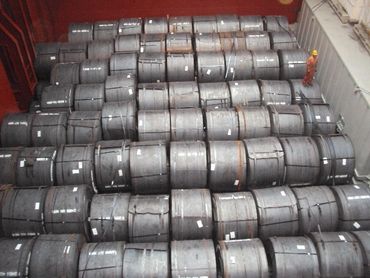 Large steel coils stacked in a warehouse with a worker for scale.