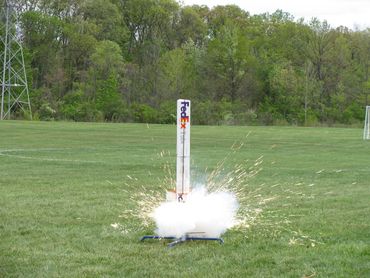 Model rocket launching on a grassy field with sparks and smoke.