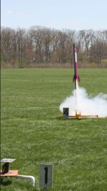 A model rocket launching from a grassy field with smoke at base.