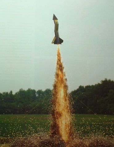 Model rocket launching with bright sparks against a natural backdrop.