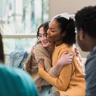 Two women warmly hugging during a group gathering indoors.