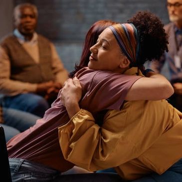 Two women share a comforting hug in a support group setting.