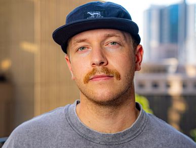 Man with a mustache wearing a navy cap and gray sweatshirt outdoors.
