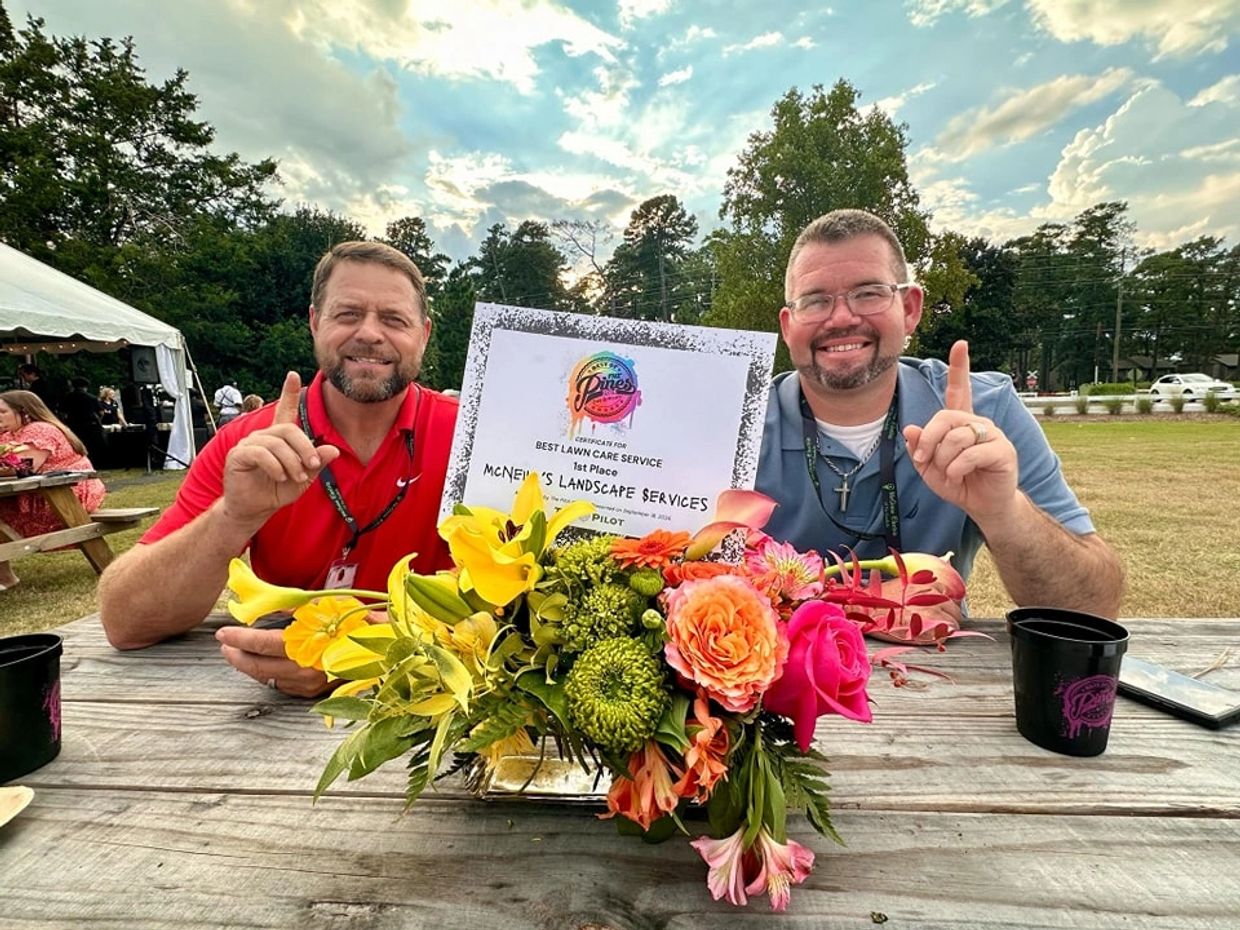 Two men proudly display a first-place award for lawn care service at an outdoor event.