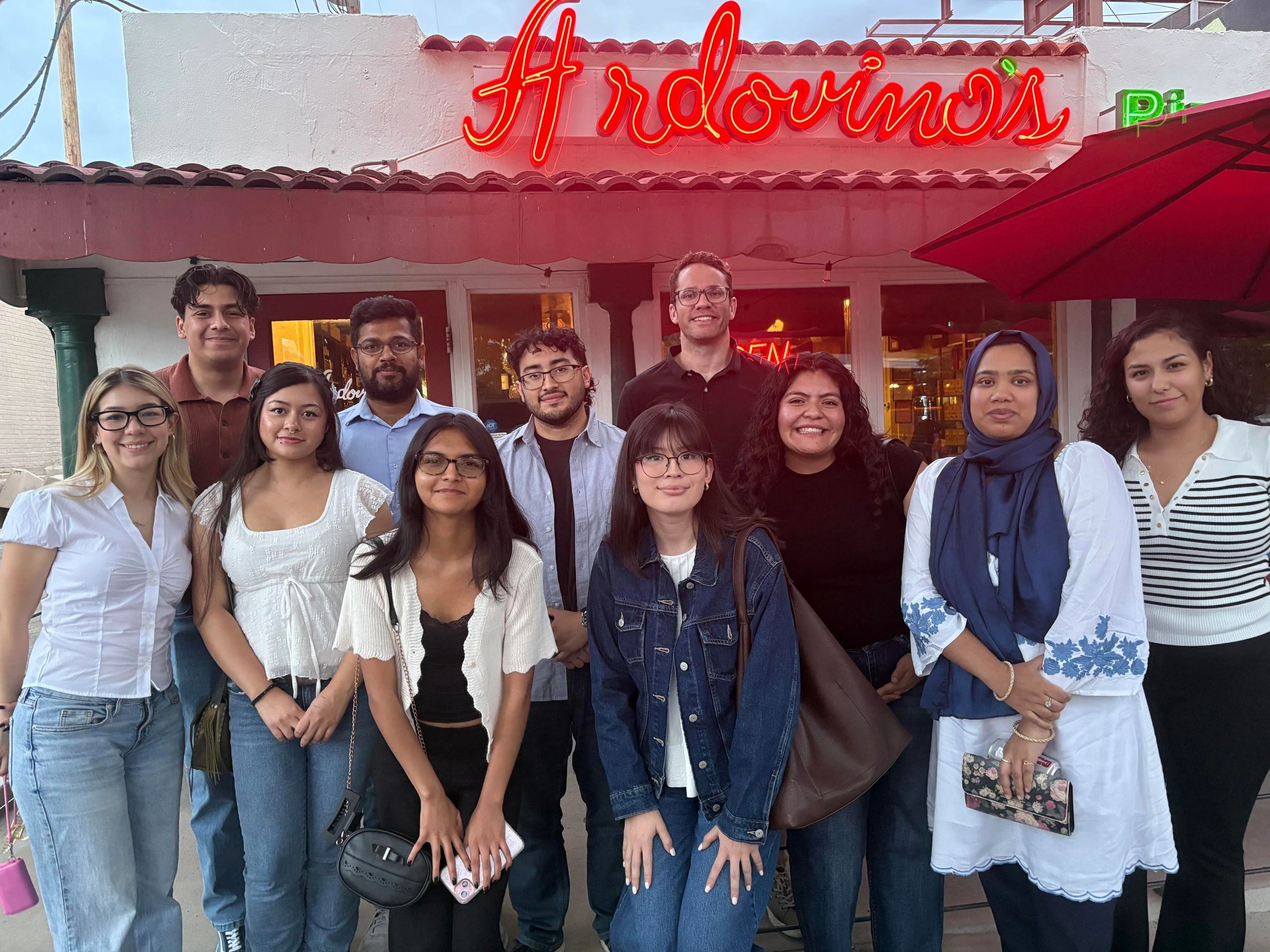 Group of diverse young people posing outside a restaurant with a neon sign.
