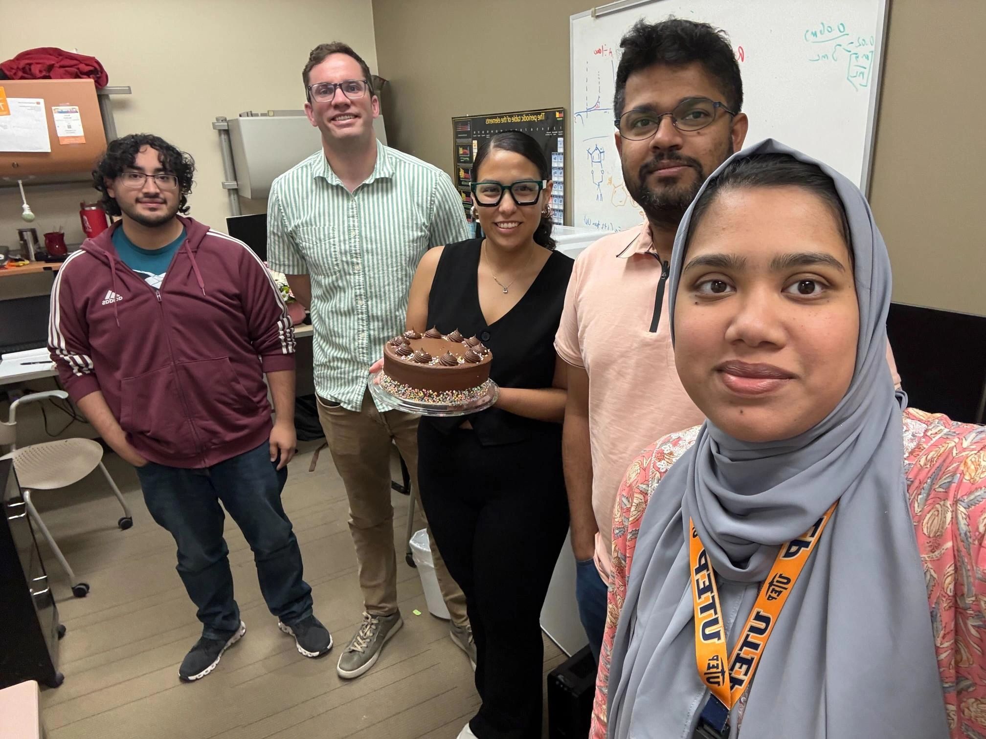 A group of five people smiling indoors, one holding a chocolate cake.