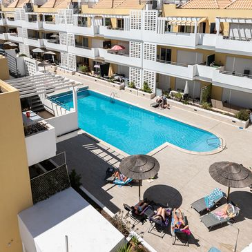 Sunny day at an apartment complex pool with people lounging under umbrellas.