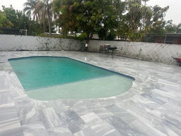 A backyard pool with gray tiled deck and surrounding greenery.