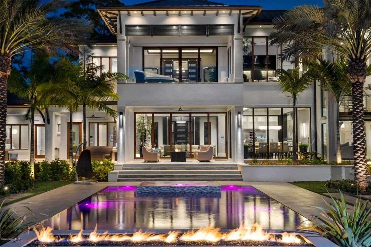 Modern luxury home with illuminated pool and fire pit at night.