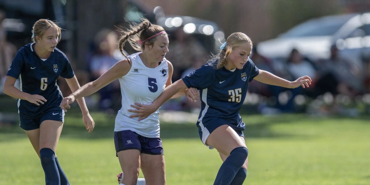 Three female soccer players compete intensely for the ball on a sunny field.