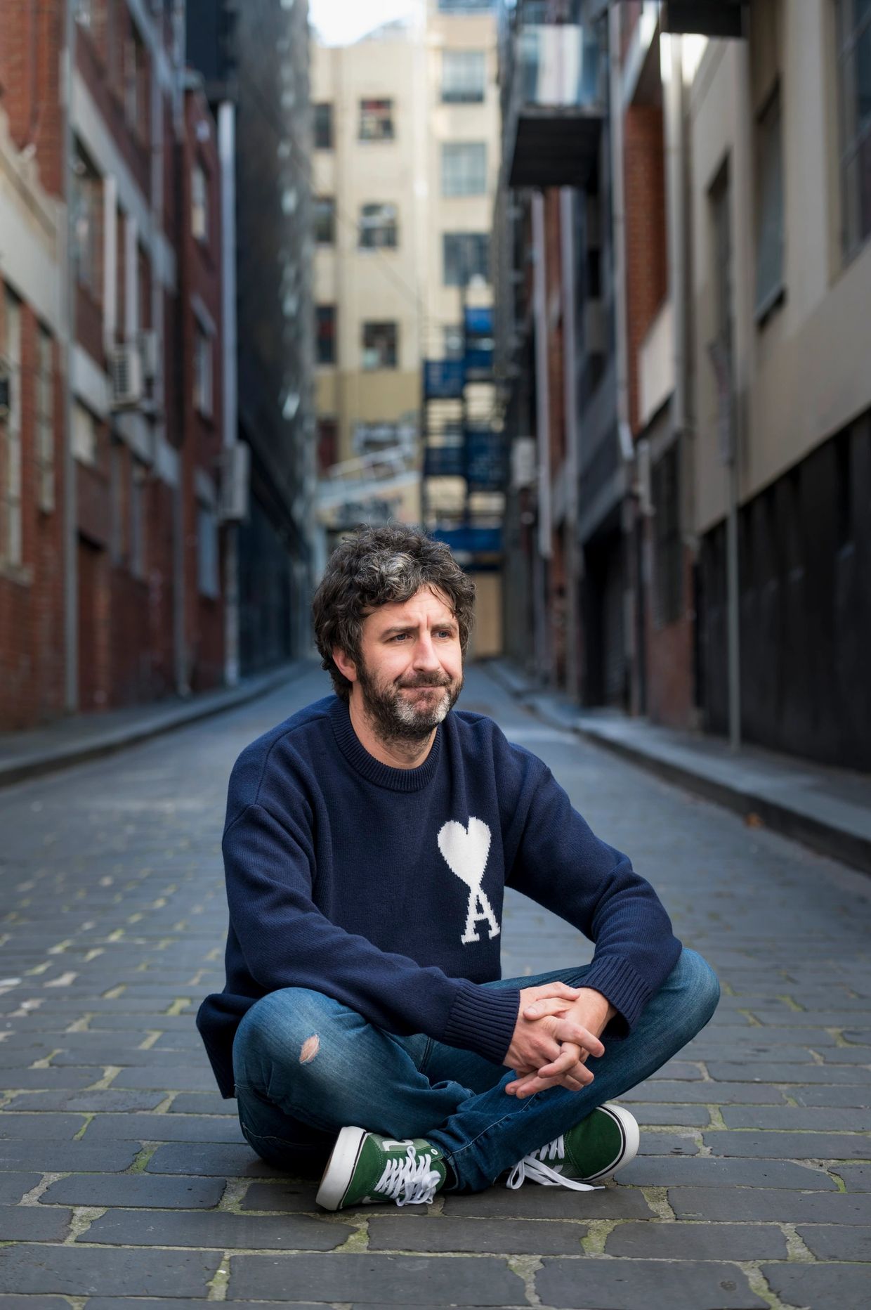 Man in a blue sweater sitting cross-legged on a cobblestone street.