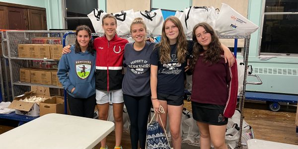 Five young women stand together smiling in a room with packed boxes and bags.