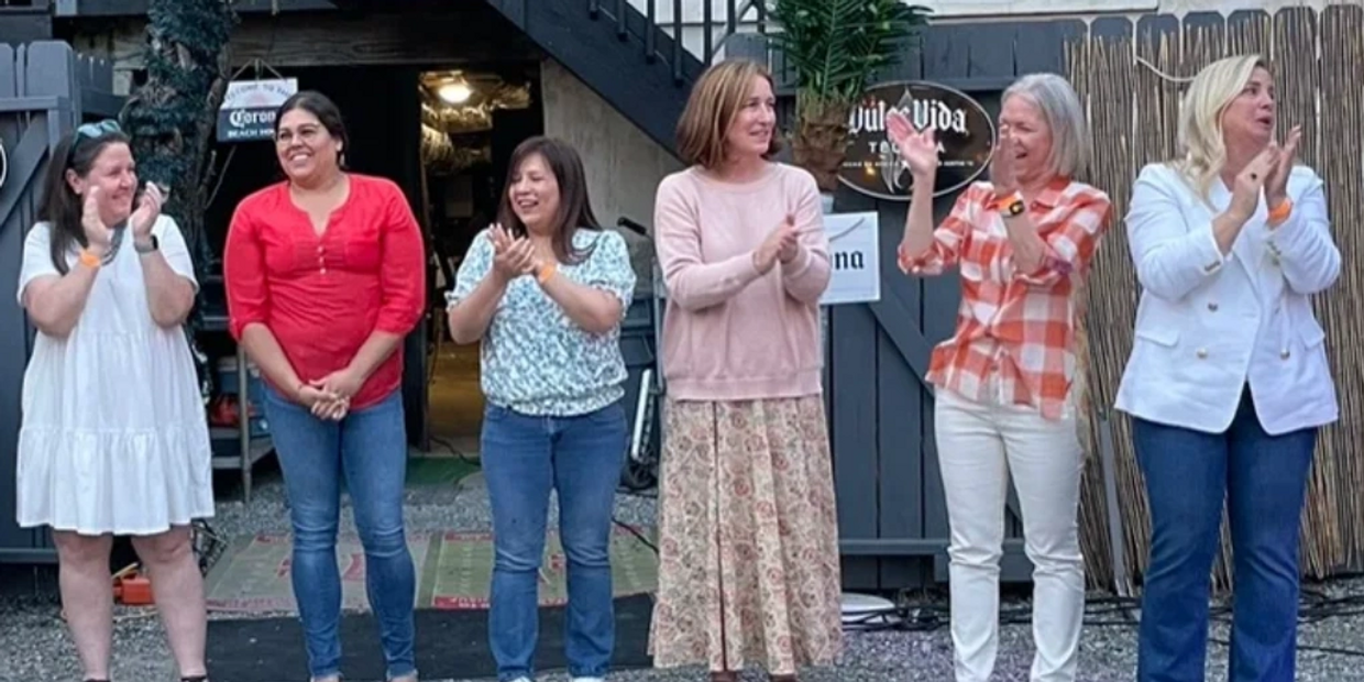 Six women stand in a row, smiling and clapping outdoors in casual attire.