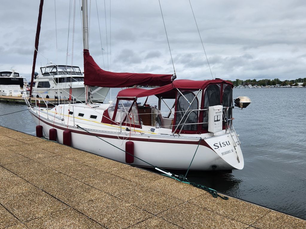 A white sailboat named Sisu docked in a marina with maroon covers on an overcast day.