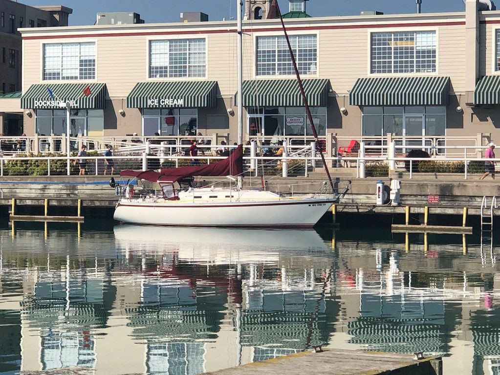Sailboat docked in calm marina waters with buildings and striped awnings reflected.