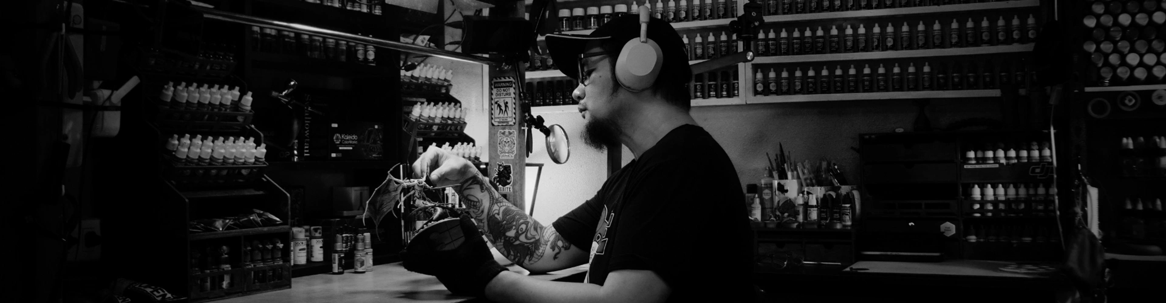 Man with headphones painting a miniature dragon in a cluttered workshop.