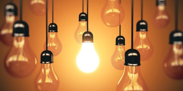 A bright light bulb glowing among many unlit bulbs against a warm background.