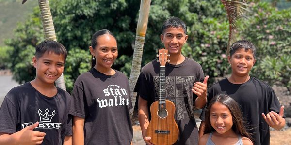 Five smiling kids posing outdoors with ukulele and palm trees.