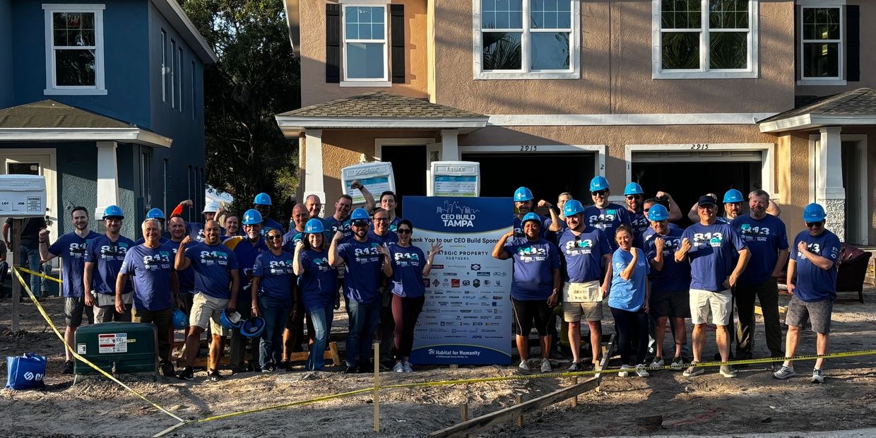 Group of volunteers in blue shirts and helmets posing in front of a Habitat for Humanity build site.