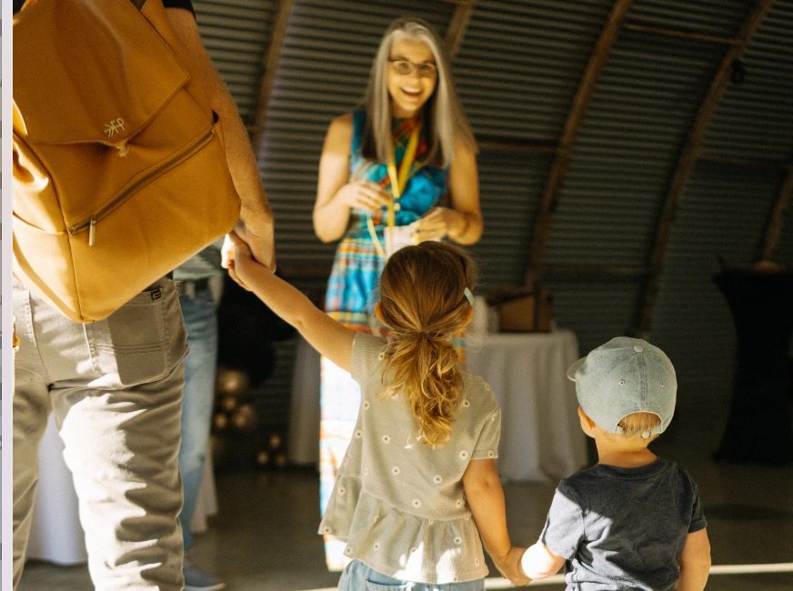 Children holding hands with an adult, facing a smiling woman indoors.
