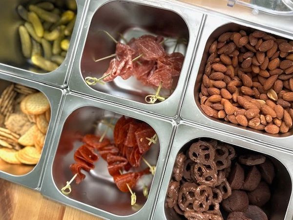 A variety of snacks including almonds, pickles, crackers, salami, pepperoni, and chocolate pretzels.