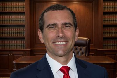 A man in a suit and red tie smiling in a wood-paneled office with shelves of books.