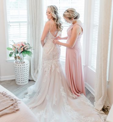 Bride getting ready with help from bridesmaid in soft-lit room.
