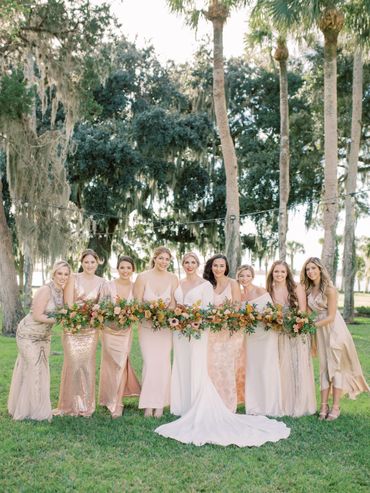 Bride and bridesmaids in elegant dresses holding long floral bouquets outdoors.