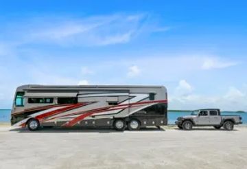 A large RV being towed by a pickup truck near the ocean under a clear blue sky.