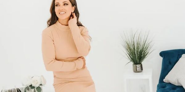 A woman in a beige dress sitting and smiling in a bright room.