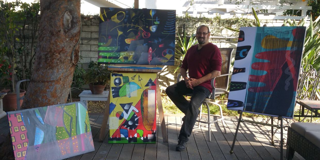 Man sitting with colorful abstract paintings displayed outdoors.