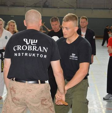 Two men practicing self-defense techniques in a martial arts class with onlookers.
